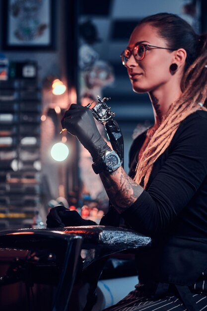 Jovem atraente tatuador em luvas pretas está ajustando sua máquina de tatuagem antes da sessão de tatuagem.