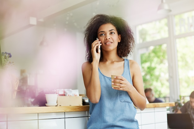 Jovem atraente universidade universitária de pele escura, com cabelos ondulados escuros na camisa azul casual, falando no telefone com o namorado, olhando de lado com uma expressão calma, tomando café relaxante após o estudo