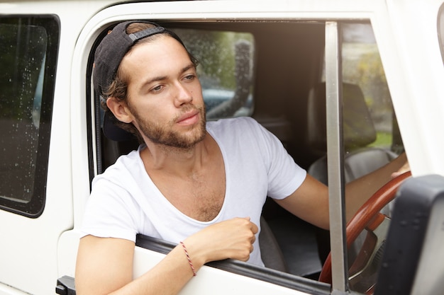 Jovem aventureiro barbudo vestindo snapback preto dirigindo seu SUV branco, indo para uma viagem de safari, enfiando a cabeça pela janela aberta, tendo um olhar cansado e infeliz