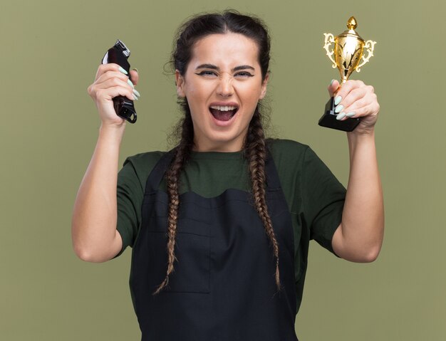 Jovem barbeira de uniforme empolgada segurando uma máquina de cortar cabelo com a taça do vencedor isolada na parede verde oliva