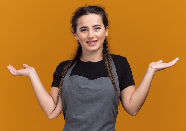 Foto grátis jovem barbeira de uniforme sorridente, espalhando as mãos isoladas na parede laranja