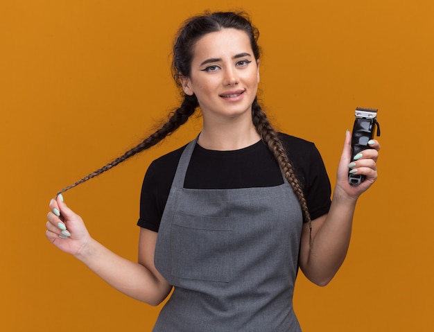 Foto grátis jovem barbeira de uniforme sorridente segurando uma máquina de cortar cabelo e segurando o cabelo isolado na parede laranja