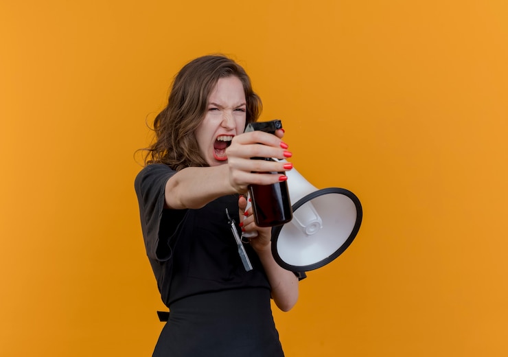Foto grátis jovem barbeira eslava usando uniforme gritando no alto-falante e olhando e esticando o frasco de spray isolado em um fundo laranja com espaço de cópia