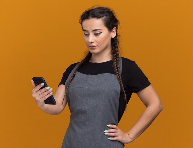 Jovem barbeira impressionada de uniforme segurando e olhando para o telefone, colocando a mão no quadril isolado na parede laranja