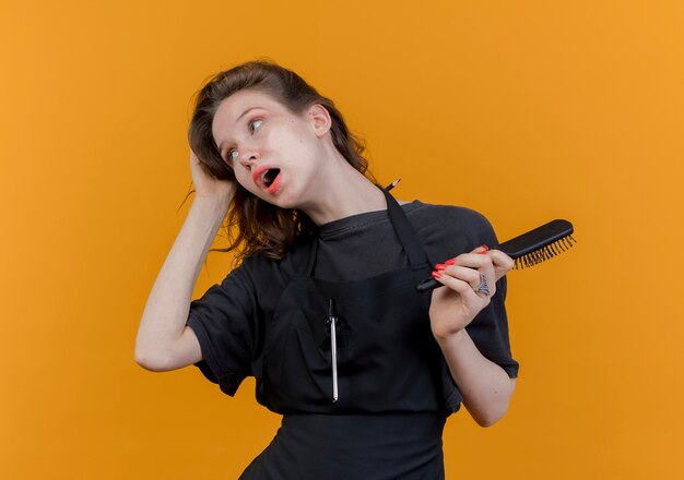 jovem barbeira impressionada vestindo uniforme olhando para o lado segurando o pente e agarrando o cabelo