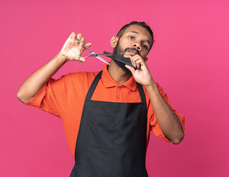Foto grátis jovem barbeiro afro-americano concentrado vestindo uniforme, olhando em linha reta, segurando uma tesoura e cortando pente modelando sua própria barba