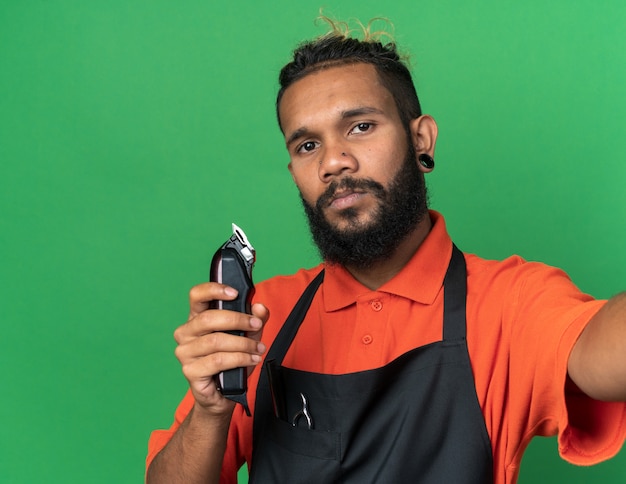 Jovem barbeiro afro-americano confiante, usando uniforme, estendendo a mão em direção à câmera, segurando uma tesoura de cabelo isolada na parede verde