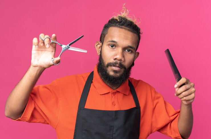 Foto grátis jovem barbeiro afro-americano confiante usando uniforme segurando uma tesoura e um pente