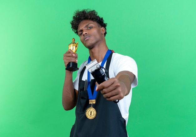 Jovem barbeiro afro-americano confiante vestindo uniforme e medalha segurando uma tesoura de cabelo para a câmera com a taça do vencedor isolada no fundo verde