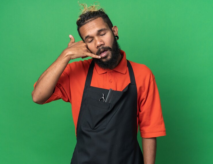 Foto grátis jovem barbeiro afro-americano de uniforme, fazendo sinal de chamada com os olhos fechados