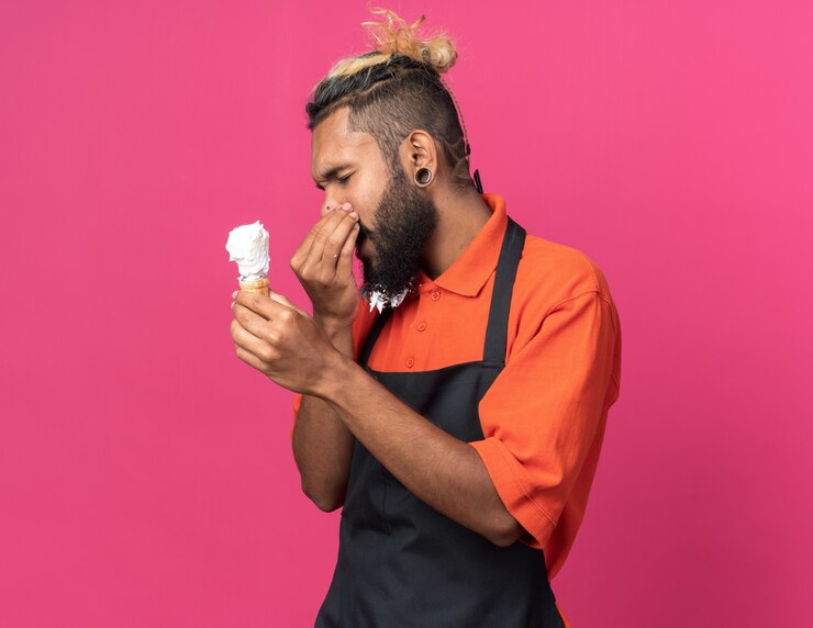 Foto grátis jovem barbeiro afro-americano enojado, usando uniforme, segurando o pincel de barbear, fazendo gesto de mau cheiro com os olhos fechados