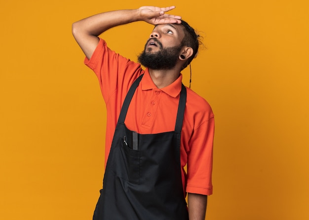 Foto grátis jovem barbeiro afro-americano impressionado, vestindo uniforme, tocando a testa, olhando para cima, isolado na parede laranja com espaço de cópia