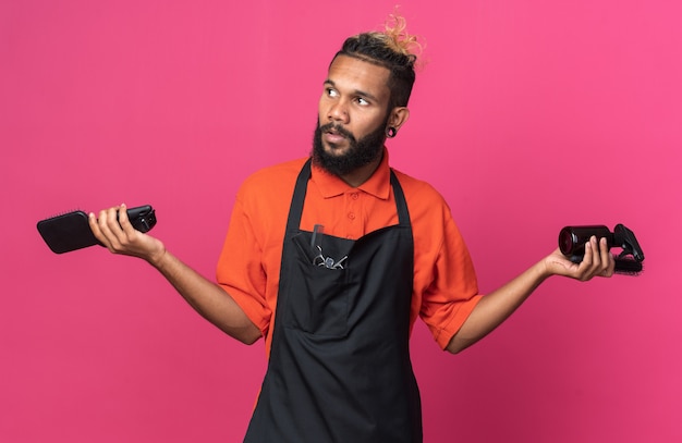 Jovem barbeiro afro-americano sem noção, usando uniforme, segurando pentes e spray de cabelo olhando para o lado