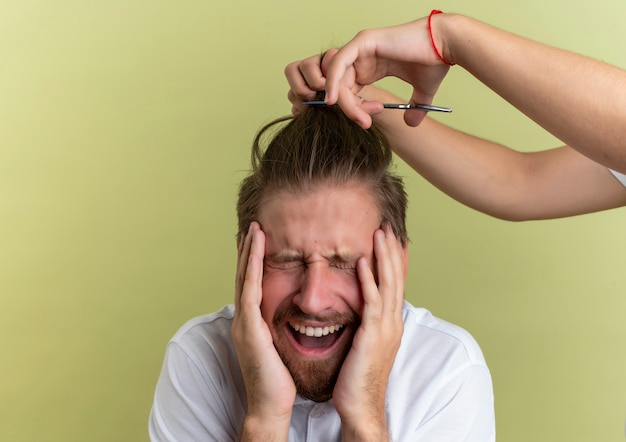 Foto grátis jovem barbeiro bonito colocando as mãos no rosto com os olhos fechados com medo de ter todo o seu cabelo cortado isolado em um fundo verde oliva com espaço de cópia