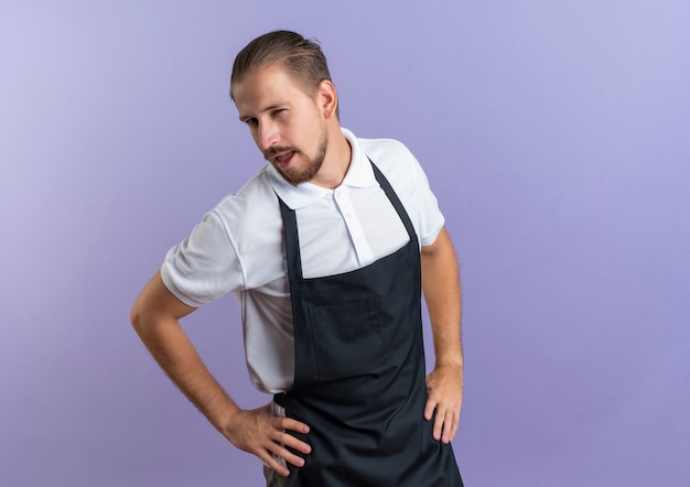 Jovem barbeiro bonito confiante vestindo uniforme colocando as mãos na cintura isolada em roxo com espaço de cópia