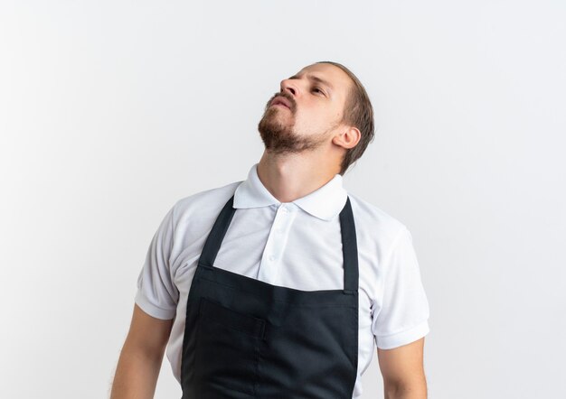 Jovem barbeiro bonito confiante vestindo uniforme, olhando para cima, isolado no branco com espaço de cópia