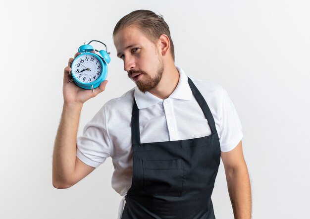 Jovem barbeiro bonito confiante vestindo uniforme segurando um despertador isolado no branco