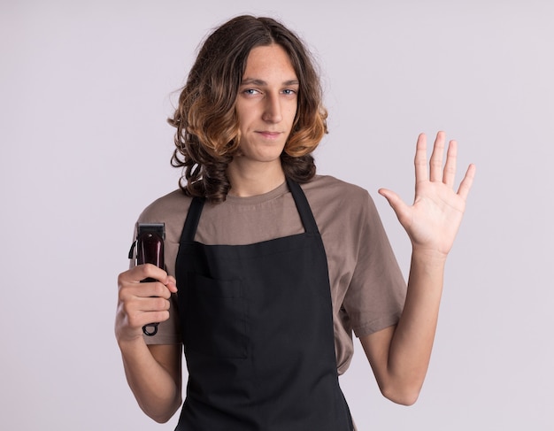 Jovem barbeiro bonito confiante vestindo uniforme segurando uma máquina de cortar cabelo, olhando para a frente, mostrando cinco com a mão isolada na parede branca
