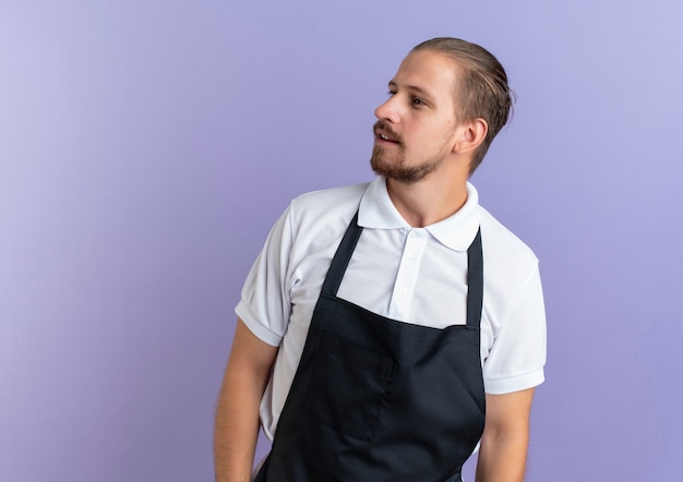 Jovem barbeiro bonito impressionado usando uniforme em pé e olhando para o lado isolado em roxo com espaço de cópia