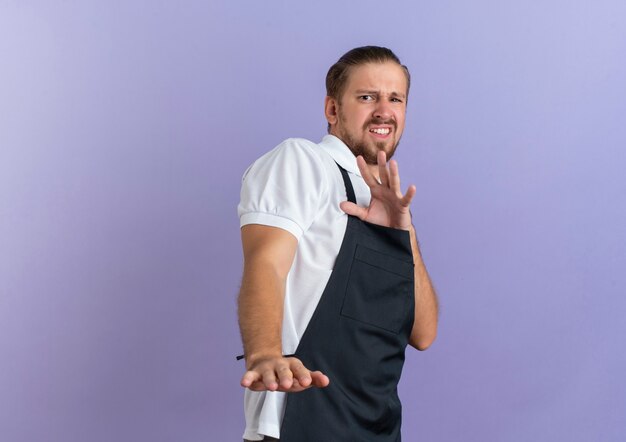 Jovem barbeiro bonito irritado, usando uniforme, estendendo a mão e gesticulando não isolado no roxo com espaço de cópia