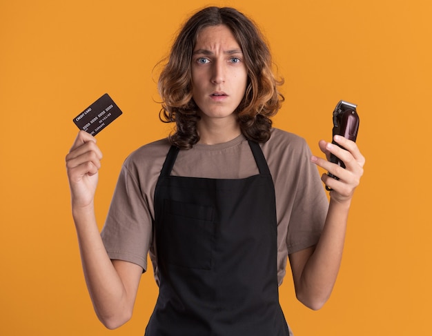 Jovem barbeiro bonito sem noção usando uniforme segurando um cartão de crédito e uma tesoura de cabelo