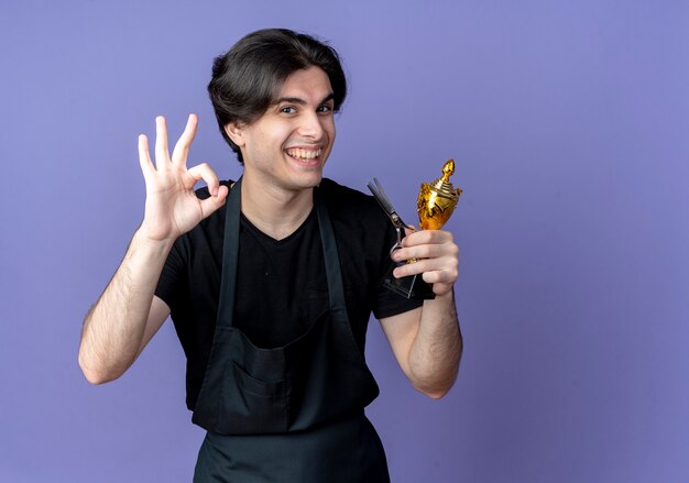 Jovem barbeiro bonito sorridente de uniforme segurando uma tesoura com a taça do vencedor e mostrando o gesto certo isolado em azul com espaço de cópia