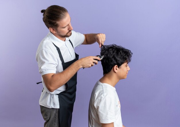 Jovem barbeiro bonito vestindo uniforme em pé em vista de perfil fazendo corte de cabelo para jovem cliente isolado em fundo roxo com espaço de cópia