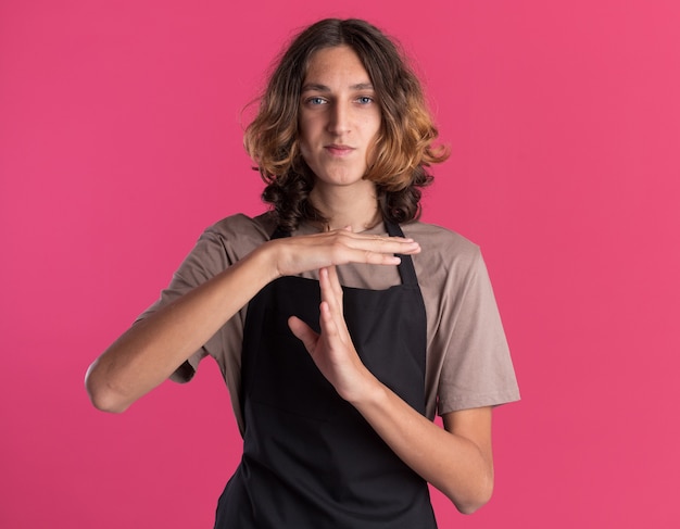 Foto grátis jovem barbeiro bonito vestindo uniforme olhando para frente fazendo gesto de tempo limite isolado na parede rosa