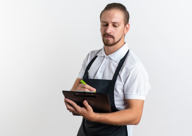 Foto grátis jovem barbeiro bonito vestindo uniforme segurando uma prancheta e uma caneta olhando para a prancheta isolada no fundo branco com espaço de cópia