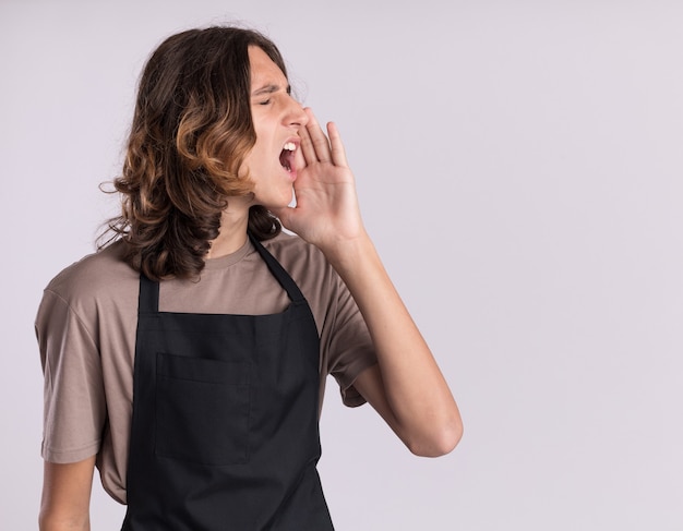 Foto grátis jovem barbeiro bonito vestindo uniforme virando a cabeça para o lado e chamando alguém de olhos fechados