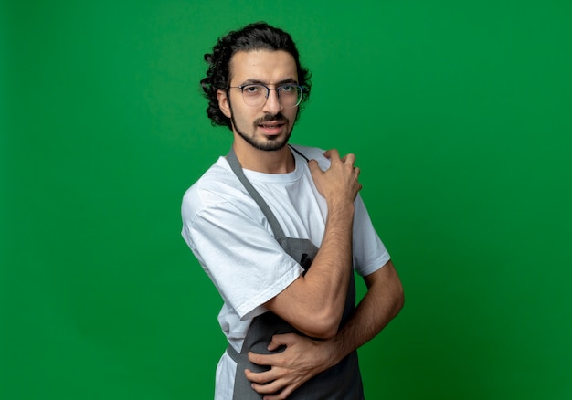 Jovem barbeiro caucasiano confiante usando óculos e faixa de cabelo ondulado em uniforme, colocando as mãos nos ombros e rins, isolado em um fundo verde com espaço de cópia