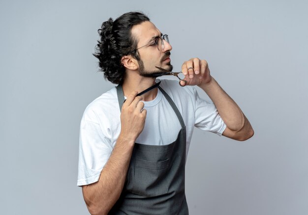 Jovem barbeiro caucasiano descontente usando óculos e faixa de cabelo ondulado em uniforme cortando e penteando a barba olhando para o lado