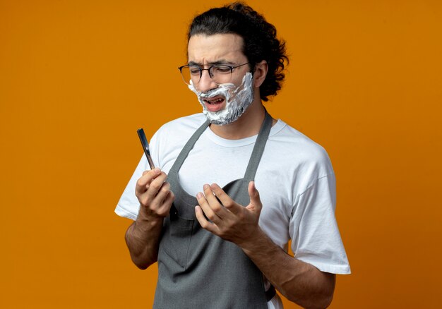 Jovem barbeiro caucasiano, irritado, usando óculos e faixa de cabelo ondulado em uniforme segurando e olhando para navalha com creme de barbear colocado no rosto mantendo as mãos no ar