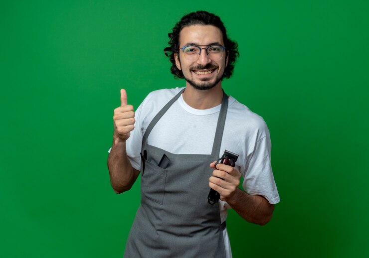 Foto grátis jovem barbeiro caucasiano sorridente, usando óculos e faixa de cabelo ondulado de uniforme, segurando uma tesoura de cabelo e aparecendo o polegar isolado em um fundo verde com espaço de cópia