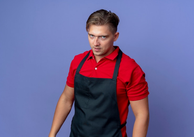 Foto grátis jovem barbeiro loira confiante de uniforme olhando para a câmera isolada no espaço violeta com espaço de cópia