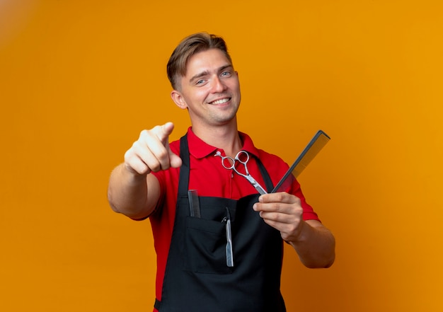 Jovem barbeiro loira sorridente com uniforme aponta para a câmera segurando uma tesoura e um pente isolado em um espaço laranja com espaço de cópia