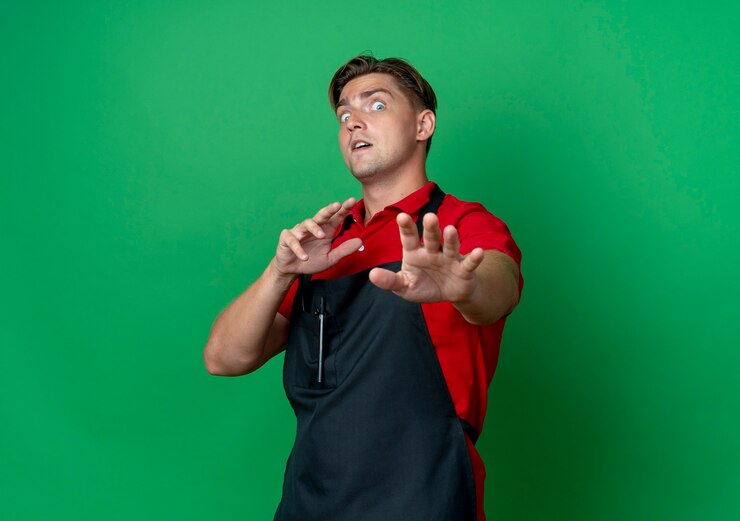 Foto grátis jovem barbeiro loiro com medo de uniforme, estendendo as mãos isoladas em um espaço verde com espaço de cópia