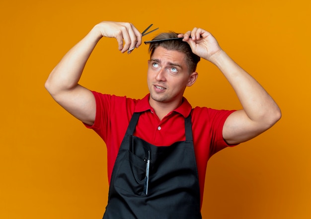 jovem barbeiro loiro de uniforme tentando cortar o cabelo com um pente e uma tesoura olhando para cima