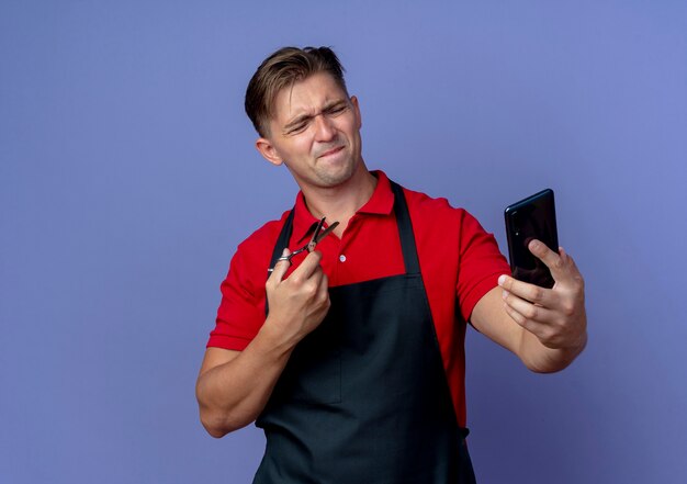 Jovem barbeiro loiro irritado de uniforme segurando uma tesoura olhando para o telefone isolado no espaço violeta com espaço de cópia