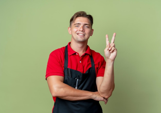 Jovem barbeiro loiro sorridente com uniforme gestos vitória mão sinal isolado no espaço verde oliva com espaço de cópia