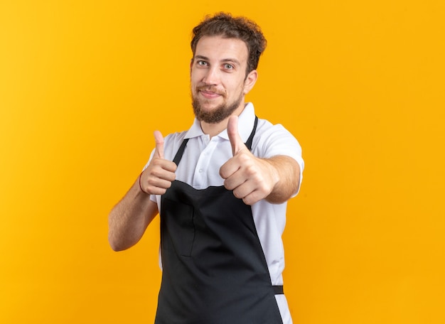 Jovem barbeiro satisfeito vestindo uniforme mostrando os polegares isolados no fundo amarelo