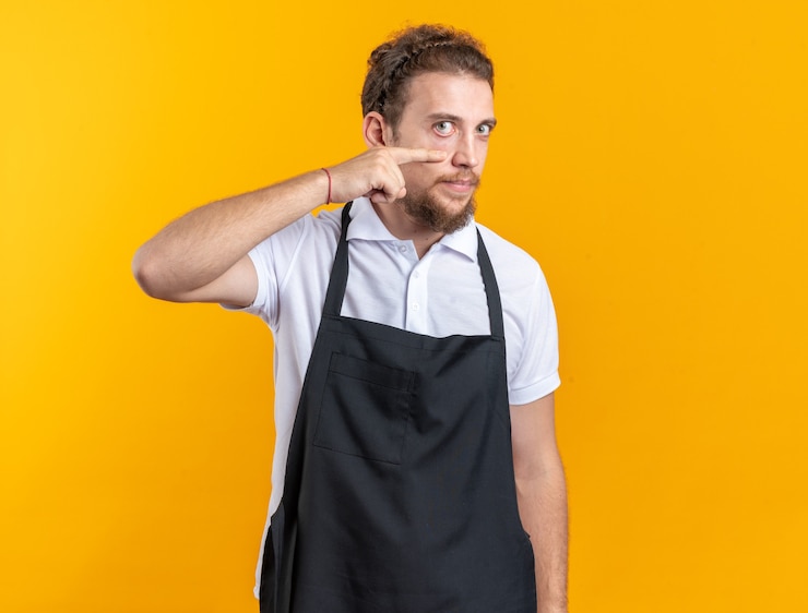 Foto grátis jovem barbeiro satisfeito vestindo uniforme puxando a pálpebra para baixo com o dedo isolado na parede amarela