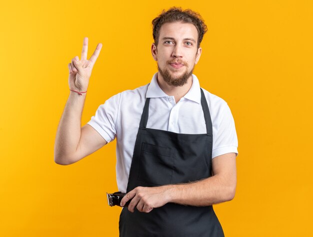 Jovem barbeiro satisfeito, vestindo uniforme, segurando uma máquina de cortar cabelo, mostrando um gesto de paz isolado na parede amarela