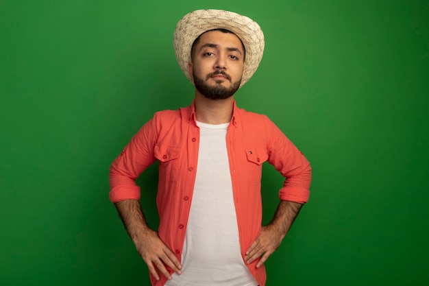 Jovem barbudo com camisa laranja e chapéu de verão, olhando com expressão séria e confiante em pé sobre a parede verde