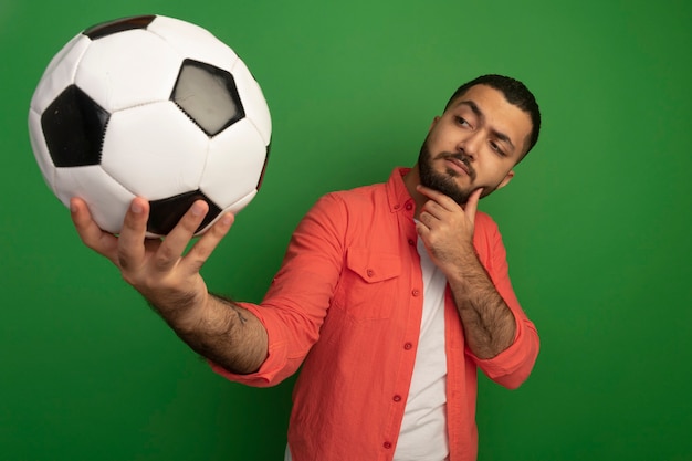 Foto grátis jovem barbudo com camisa laranja segurando uma bola de futebol olhando para ela intrigado pensando em pé sobre a parede verde