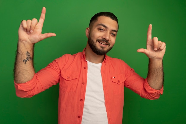 Jovem barbudo com camisa laranja sorrindo e mostrando os dedos indicadores em pé sobre a parede verde