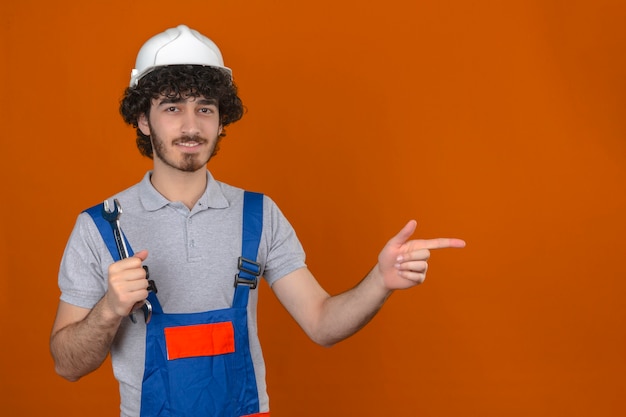 Jovem barbudo construtor bonito vestindo uniforme de construção e capacete de segurança, segurando a chave na mão, apontando para o lado enquanto sorrindo sobre parede laranja isolada