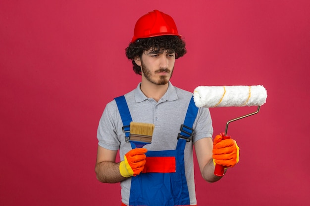 Jovem barbudo construtor bonito vestindo uniforme de construção e capacete de segurança, segurando o pincel e o rolo de pintura, olhando com o rosto sério, franzindo a testa sobre parede rosa isolada