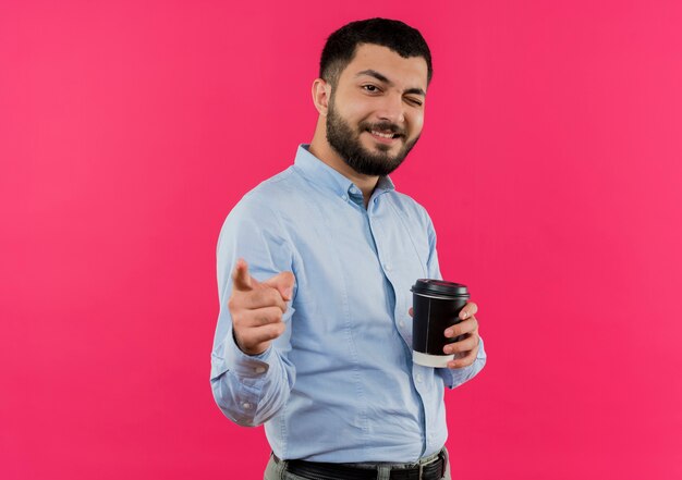 Jovem barbudo de camisa azul segurando o marcador de uma xícara de café com o dedo indicador para a câmera, sorrindo e piscando