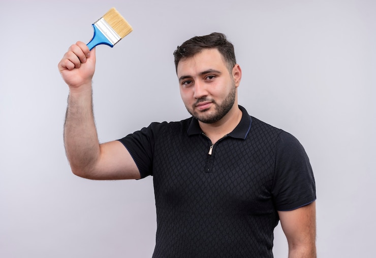 Foto grátis jovem barbudo de camisa preta segurando um pincel e levantando a mão, olhando para a câmera com um sorriso